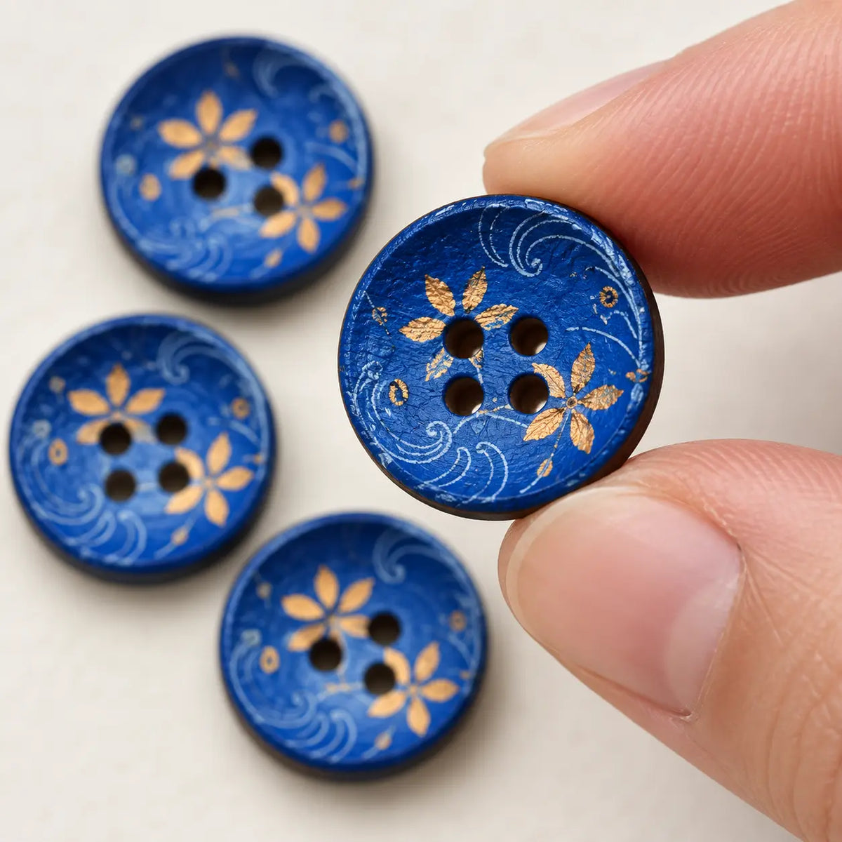 A 15 mm blue-and-gold wooden button held between fingertips with all four holes visible